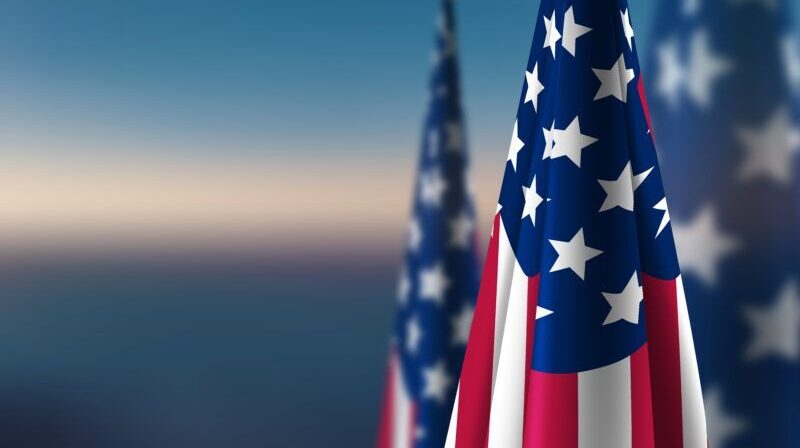 Row of American flags gathered at their poles, with one in sharp focus and a soft twilight sky in the background.
